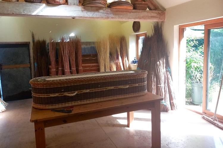 A willow coffin on a table with sheafs of willow in the background and an open door letting in sunlight.